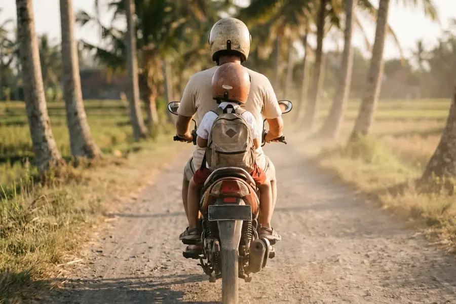 A parent and child riding a scooter along a palm-lined lane in Bali, both wearing helmets